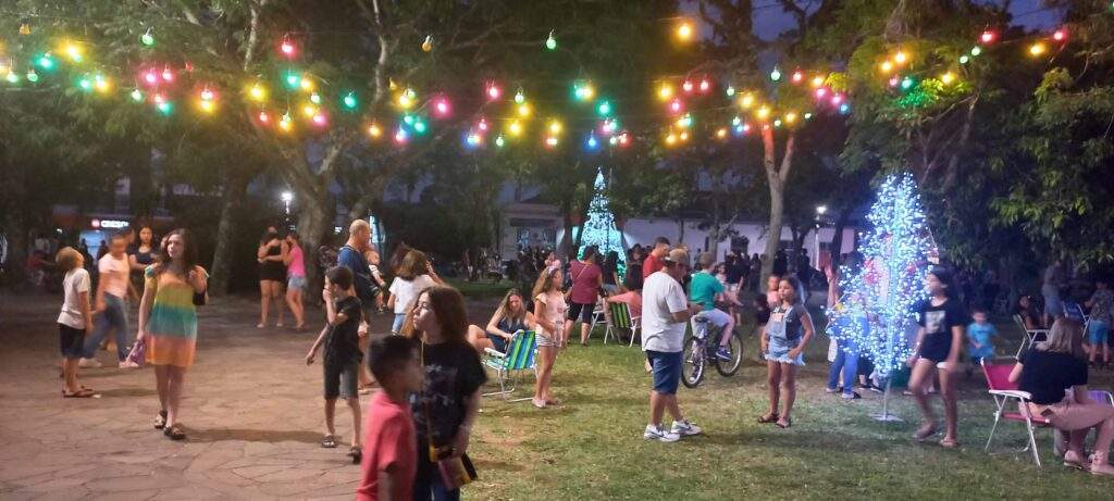 A imagem contem crianças brincando na praça sob luzes de Natal